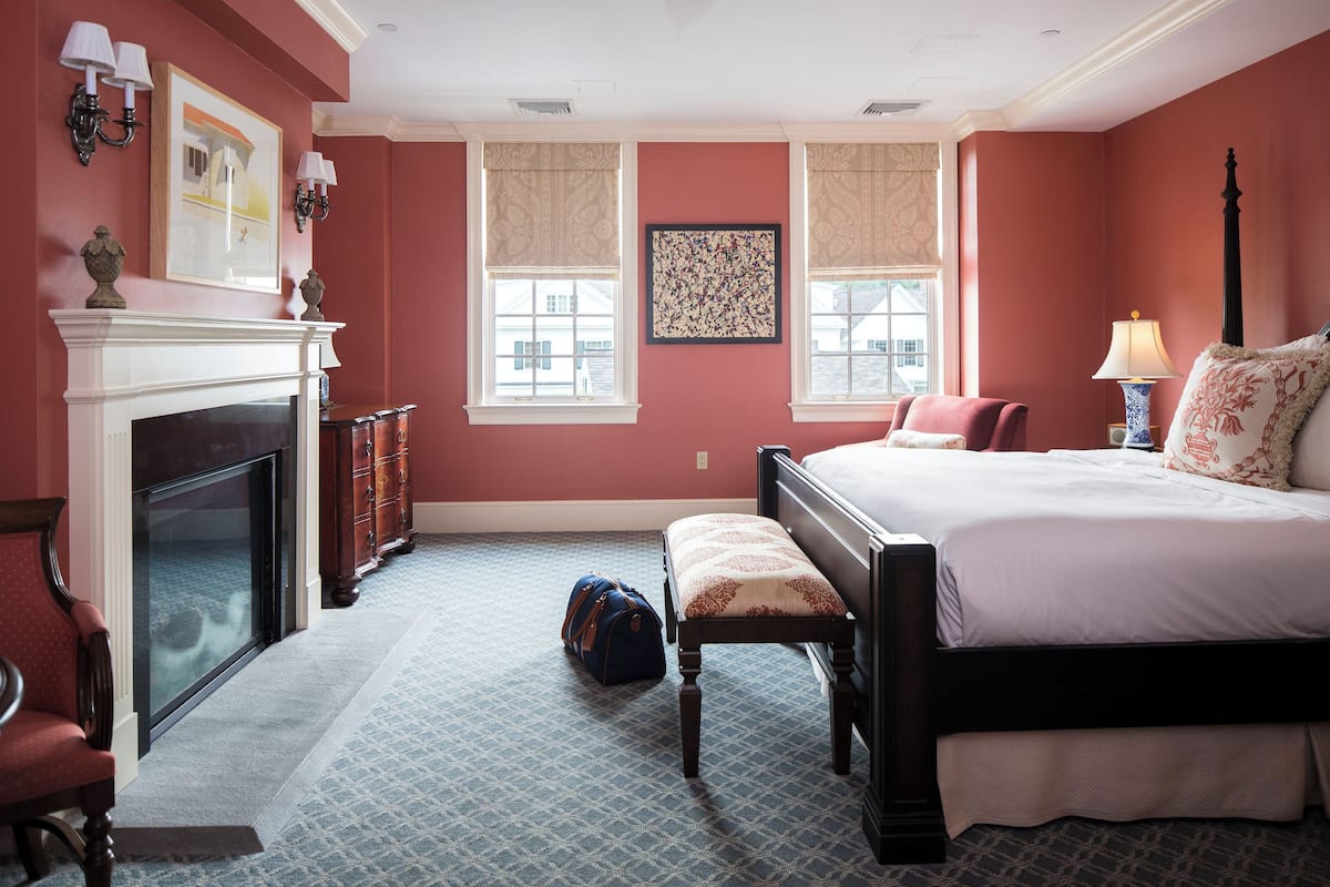 A bedroom with a large bed, a fireplace, a bench, and a suitcase on the floor.