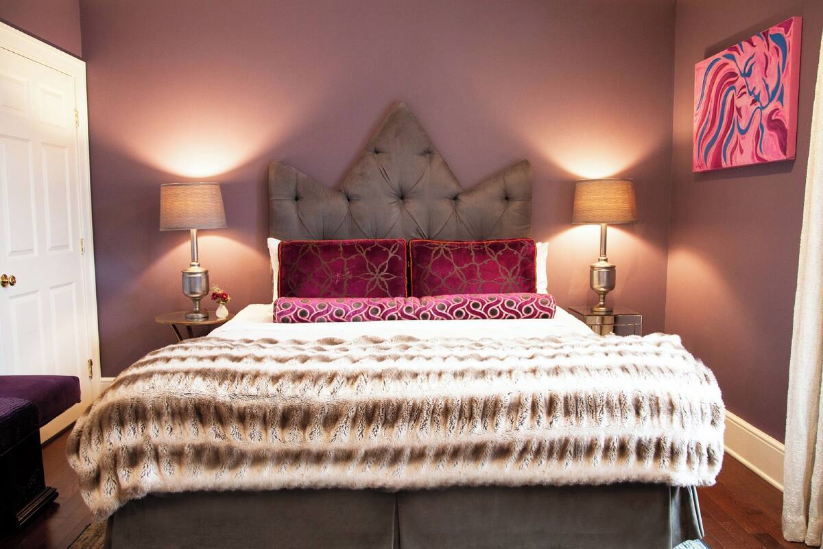 A bedroom with a tufted headboard, two bedside tables with lamps, a patterned bedspread, and a pink accent pillow.