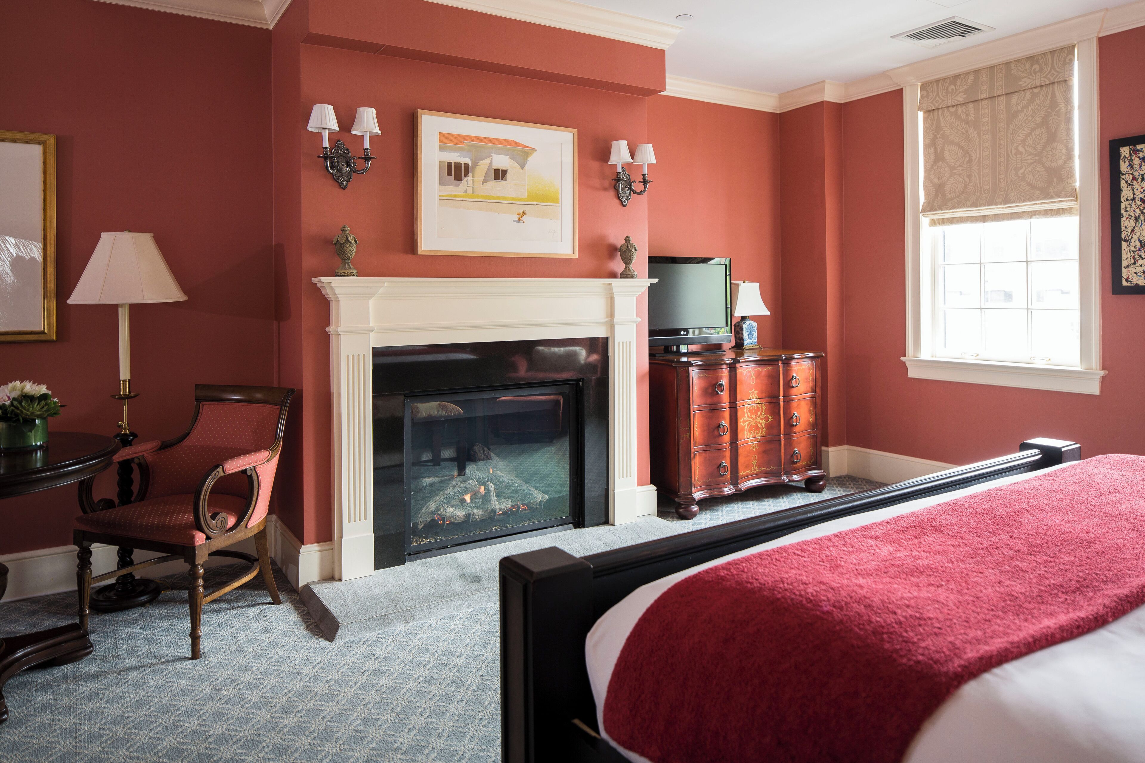 Un dormitorio con chimenea, una cama con manta roja, una silla de madera y un aparador.