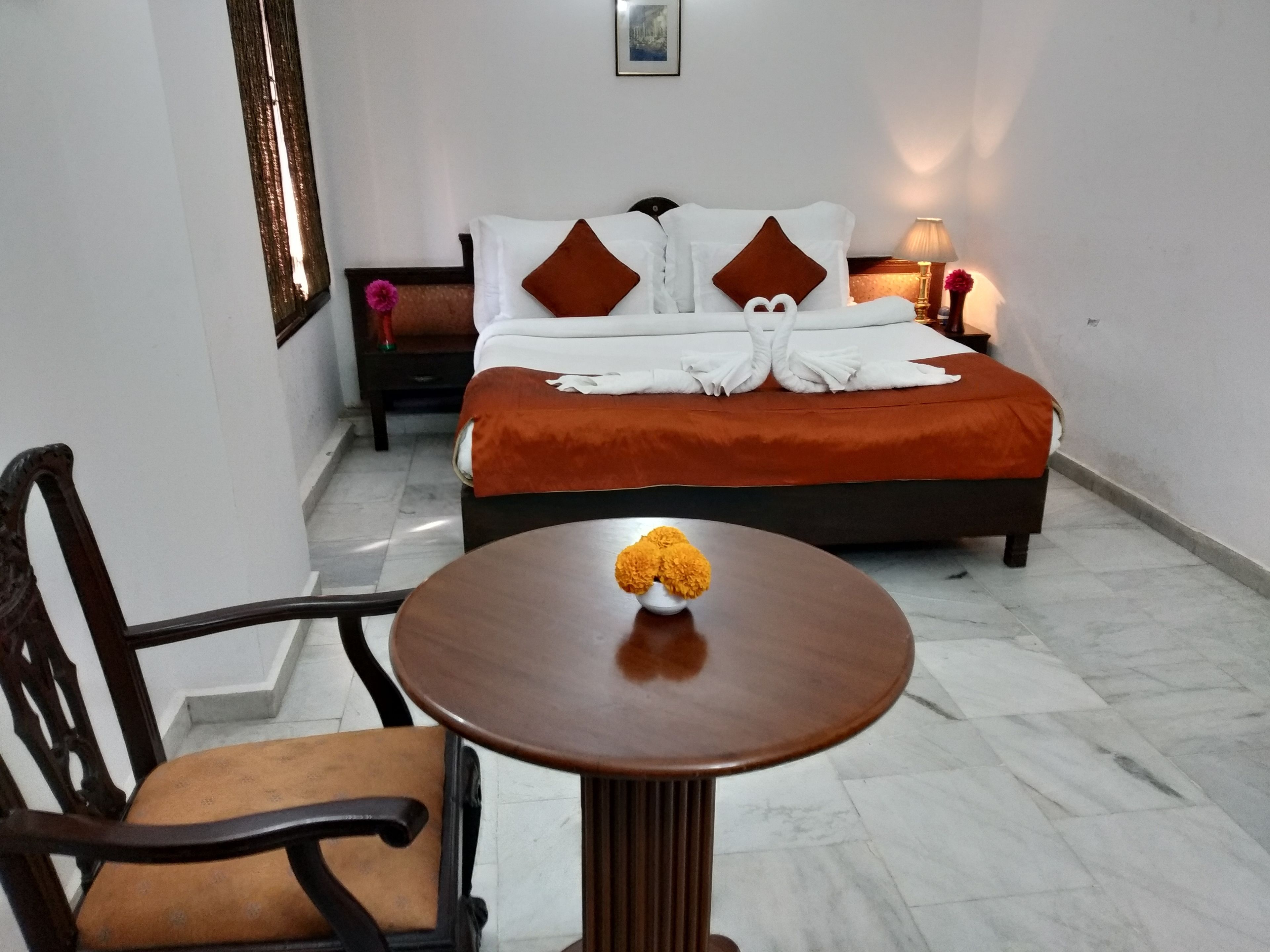 A hotel room with a bed, a chair, a table with a flower, and a nightstand with a lamp.
