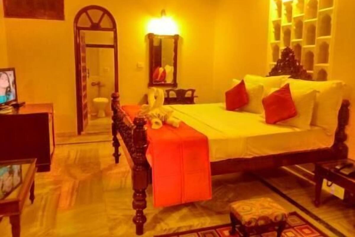 A room with a bed, a wooden chair, a small table, a mirror, and a shelf with decorative items.