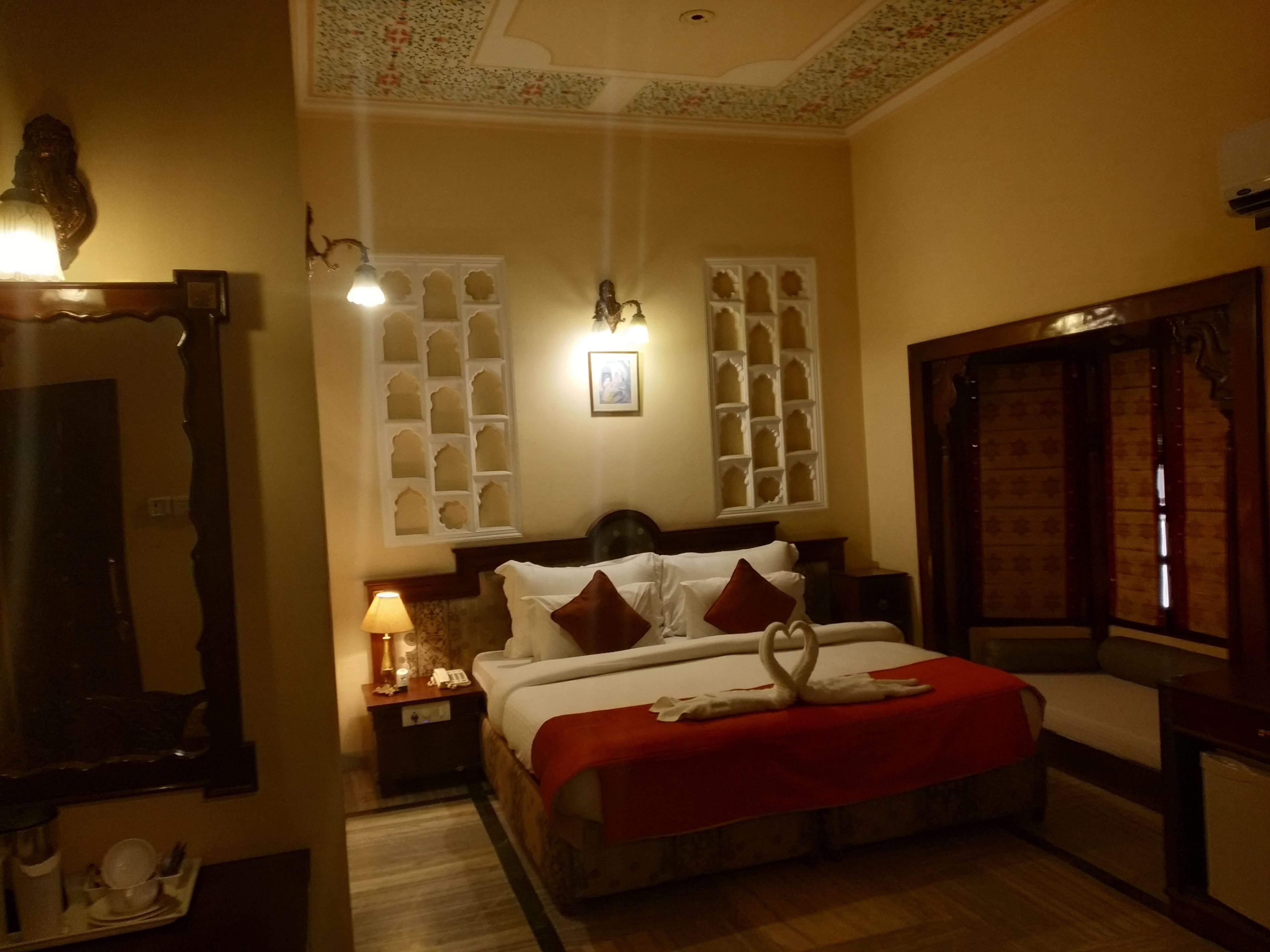 A hotel room with a bed, a nightstand, a lamp, a mirror, and a window with decorative shutters.