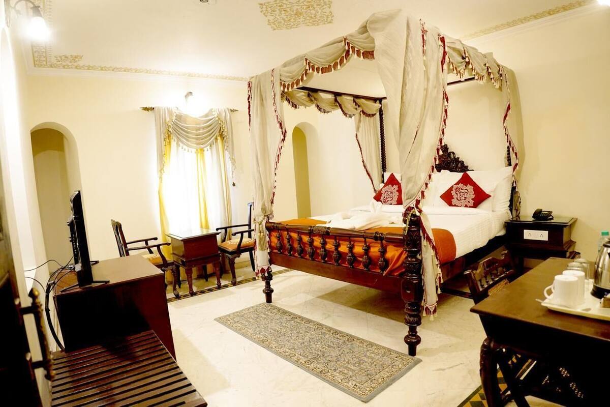 A four-poster canopy bed with a wooden frame and white bedding, a desk with a telephone, and a chair in a room with a patterned rug and arched doorway.