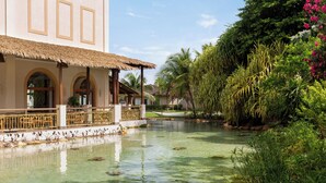 Un tranquilo patio de hotel con un estanque cristalino, abundante vegetación y un edificio tradicional con ventanas de arco y techo de paja.