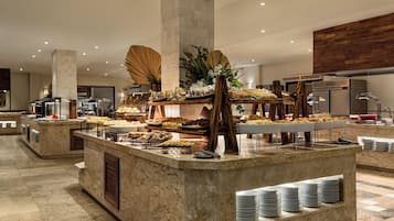 Uma área de buffet com diversas estações de comida, uma planta decorativa central e uma cozinha moderna ao fundo.
