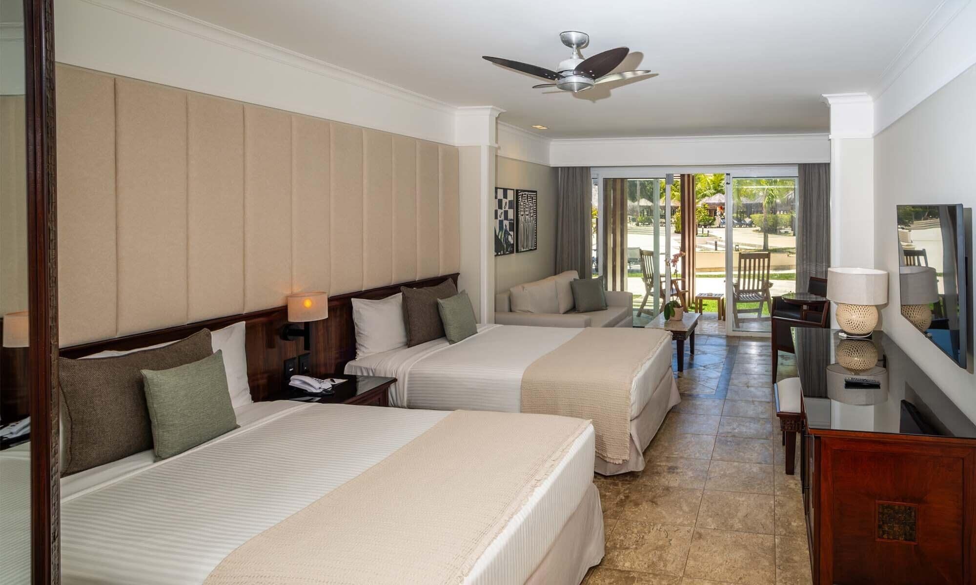 A hotel room with two beds, a ceiling fan, a sofa, and a view of the outdoors.