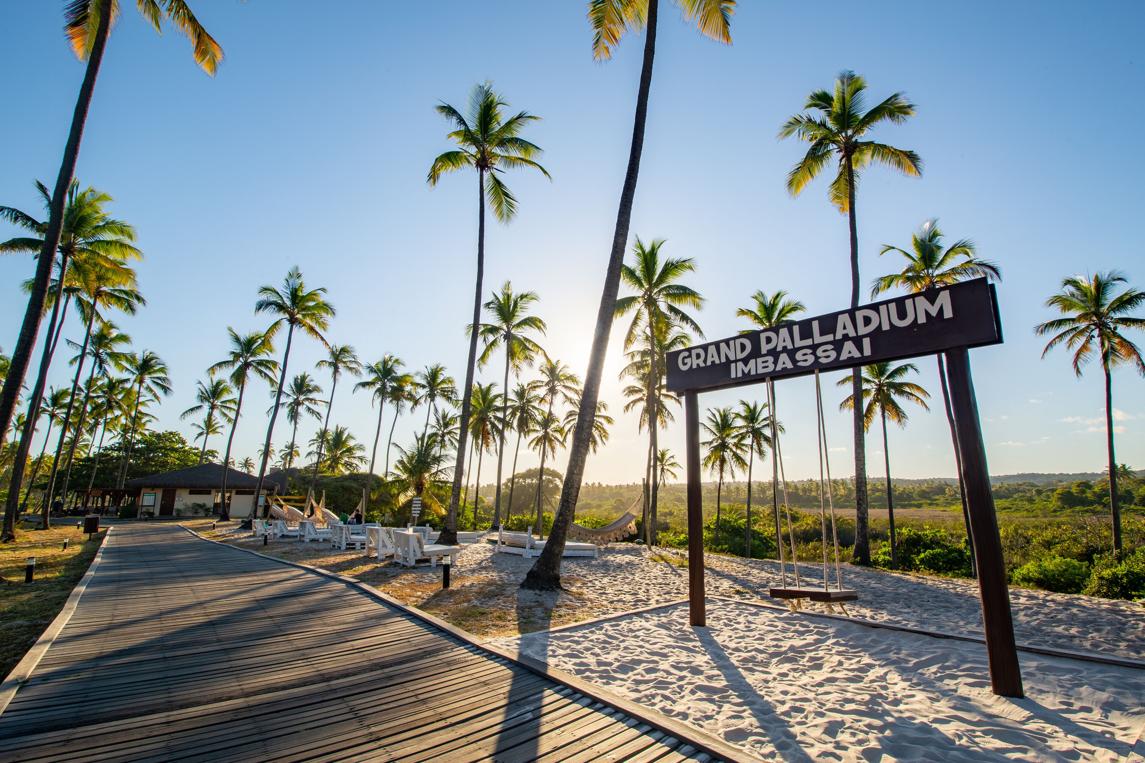 Une promenade en bois mène à une plage bordée de palmiers, où un panneau indique « Grand Palladium Imbassaï ».