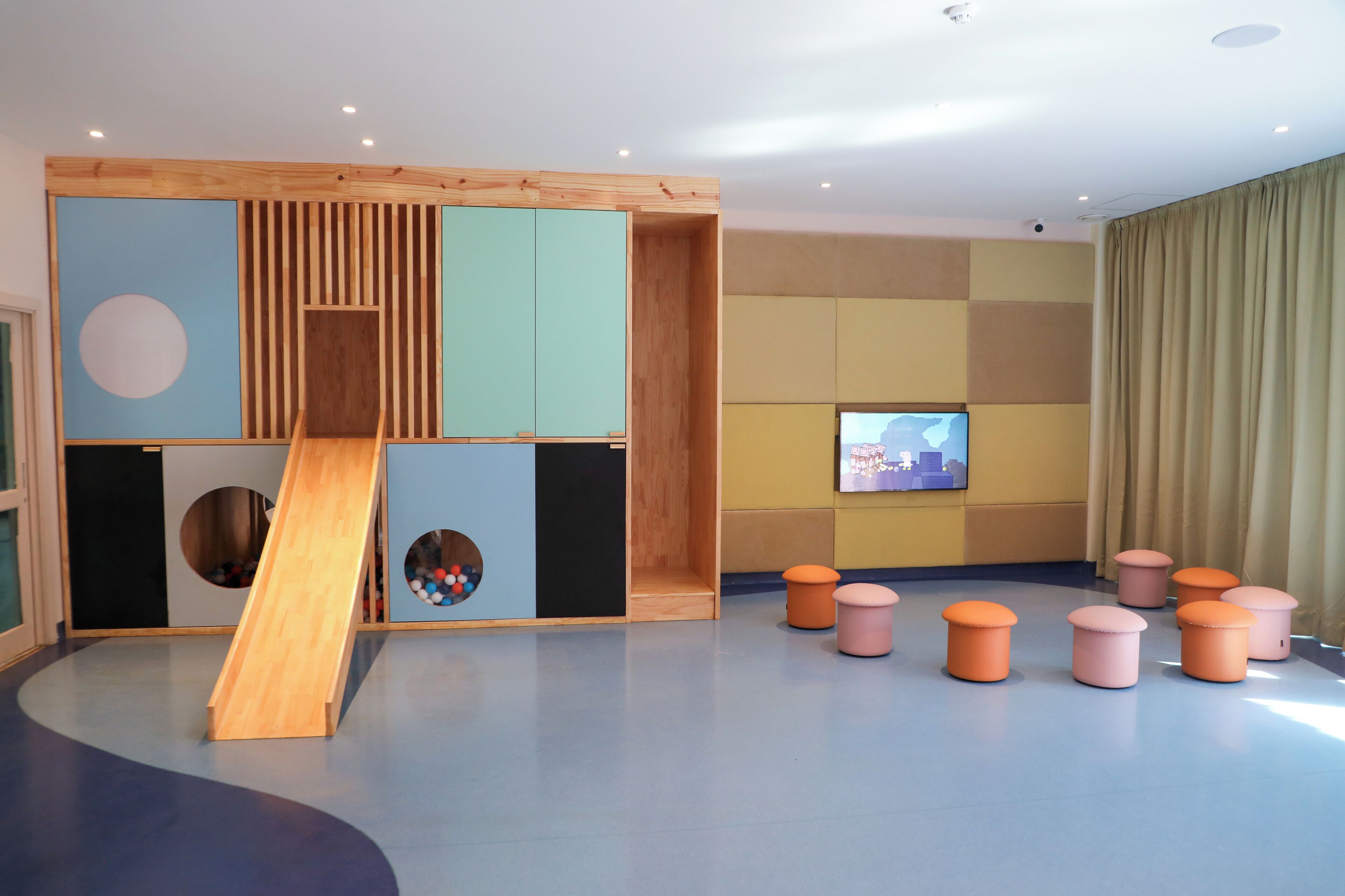 A play area with a wooden slide, colorful cushions, and a television on the wall.