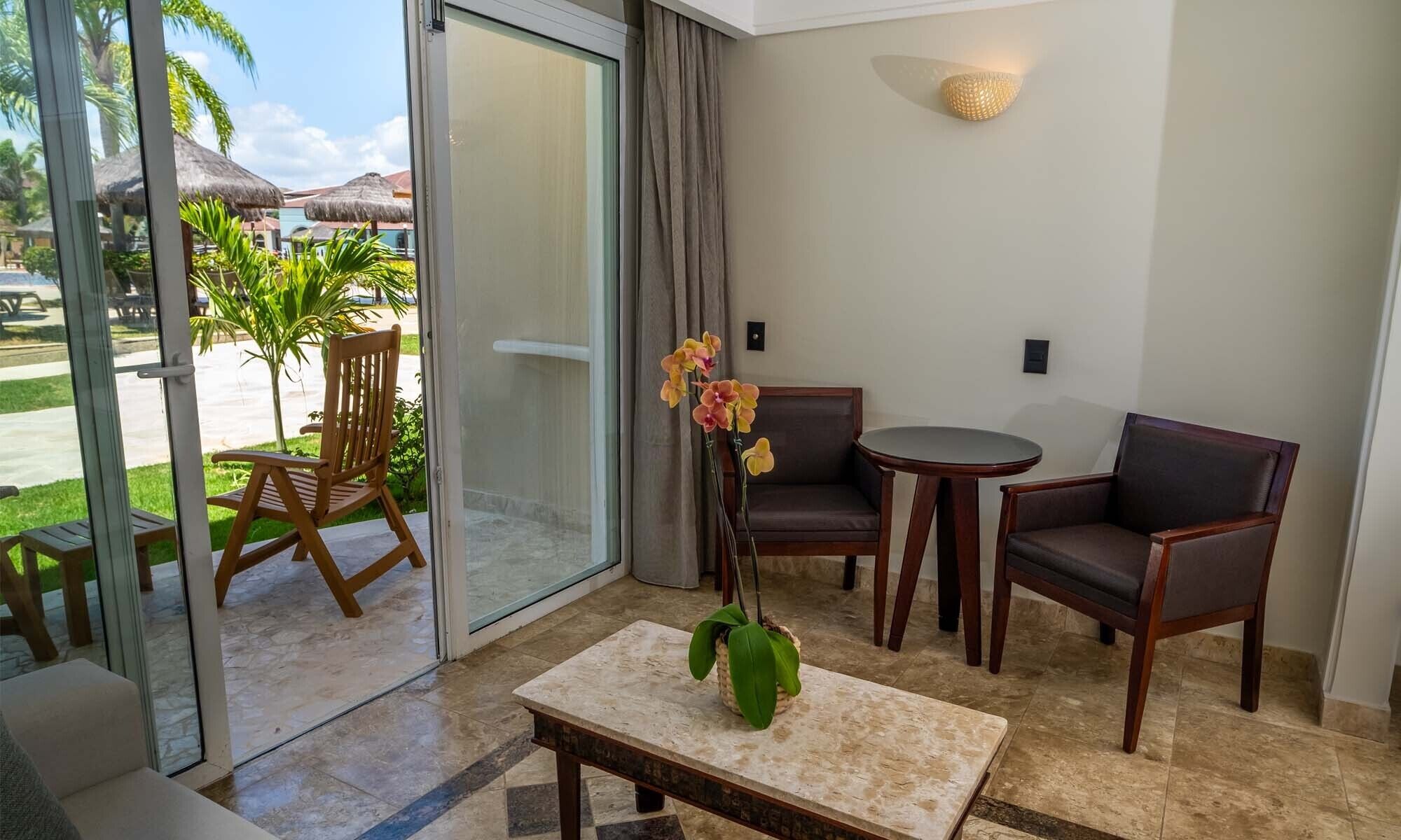 Una habitación con puerta de vidrio corrediza que da a una zona de piscina, una pequeña mesa redonda, dos sillas y una orquídea en maceta.
