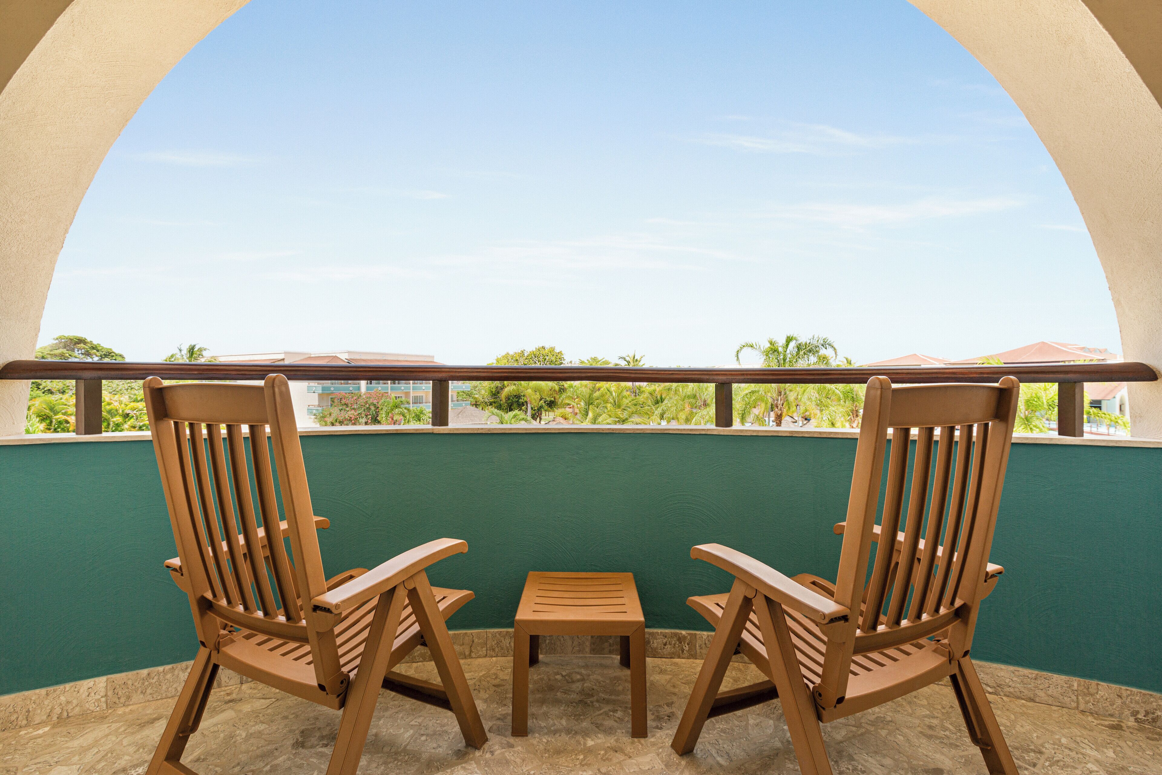 Un balcon avec deux chaises en bois et une petite table, donnant sur un espace verdoyant agrémenté d’arbres et de bâtiments.
