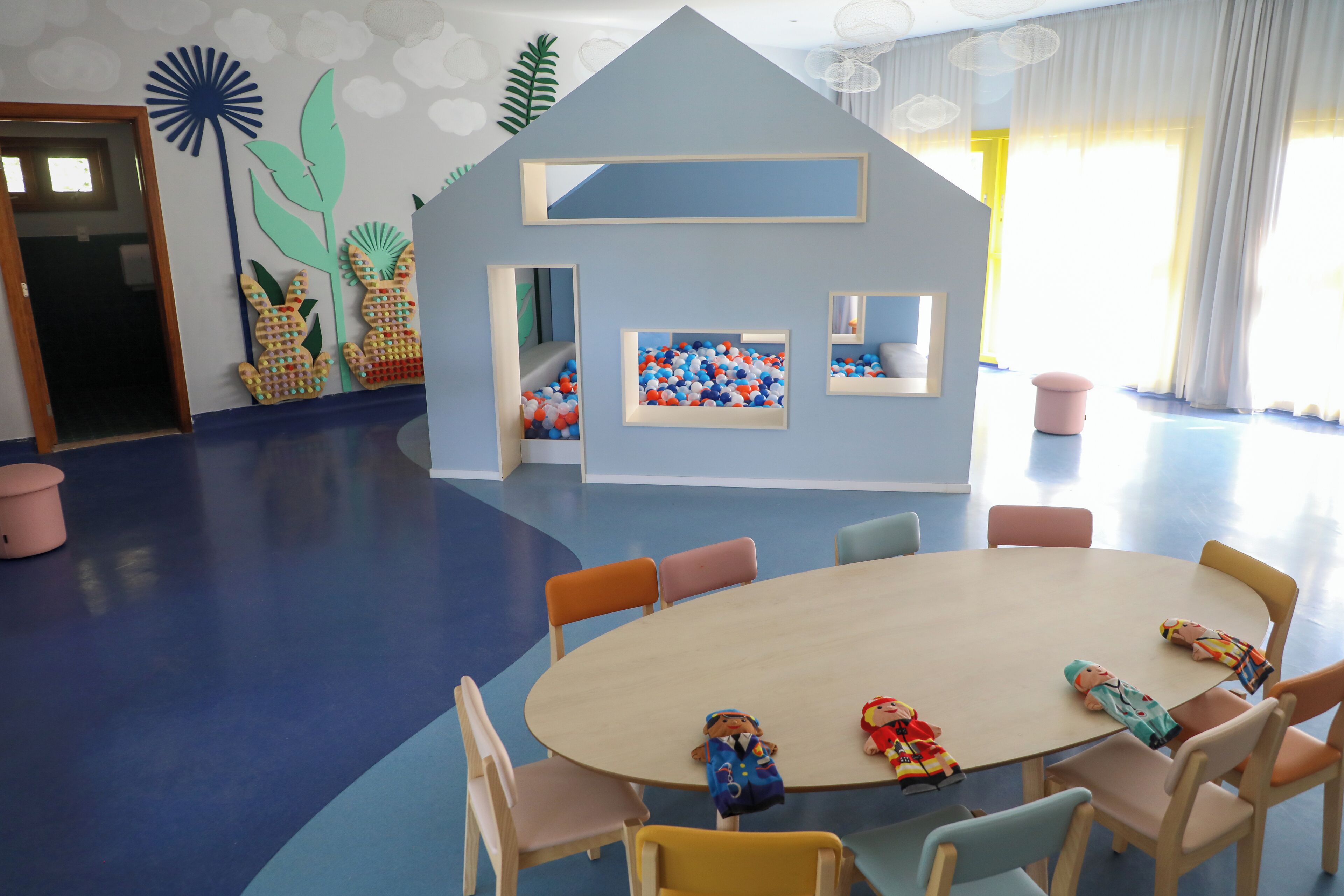 Un espace de jeux pour enfants avec une grande table ronde, des chaises colorées et une maison de jeux dotée de miroirs et d’une piscine à balles.