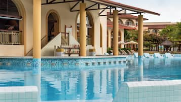 Piscina com bordas azulejadas, área de estar coberta com guarda-sóis e um edifício com janelas em arco e colunas.