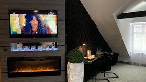 Ein modernes Wohnzimmer mit einem an der Wand montierten Flachbildfernseher, einem Kamin, einem Essbereich mit Tisch und Stühlen sowie einem großen Fenster, das für natürliches Licht sorgt.