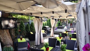 Eine Terrasse im Freien mit Tischen und Stühlen, teilweise beschattet durch Sonnenschirme, und einem an der Wand befestigten Fernseher.