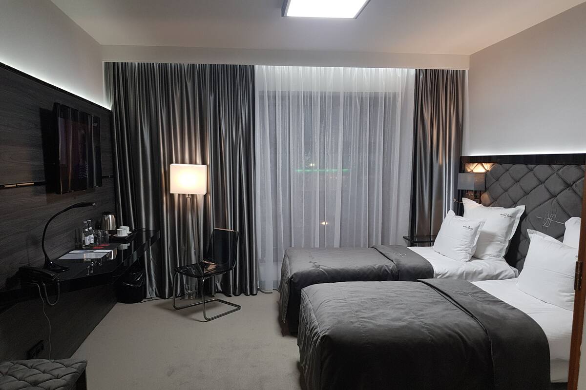Habitación de hotel con dos camas, un escritorio con lámpara, una silla y un ventanal con cortinas.