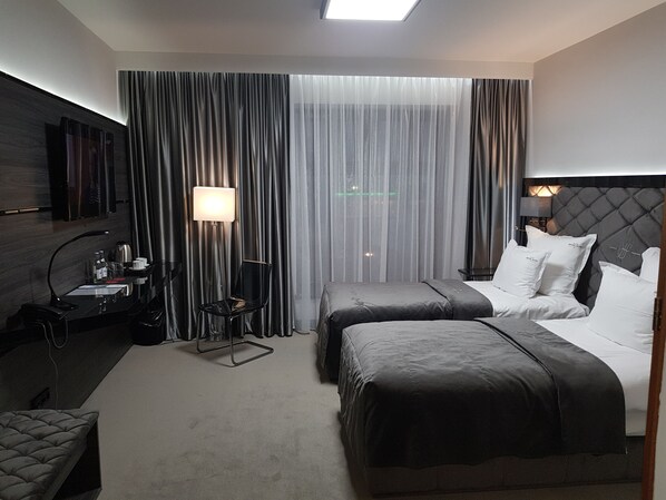 A hotel room with two beds, a desk with a lamp, a chair, and a large window with curtains.