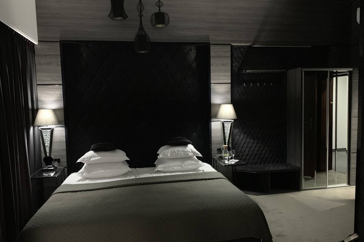 Una habitación de hotel moderna con una cama grande, dos lámparas de noche, un minibar y una zona de ducha con mampara de vidrio.