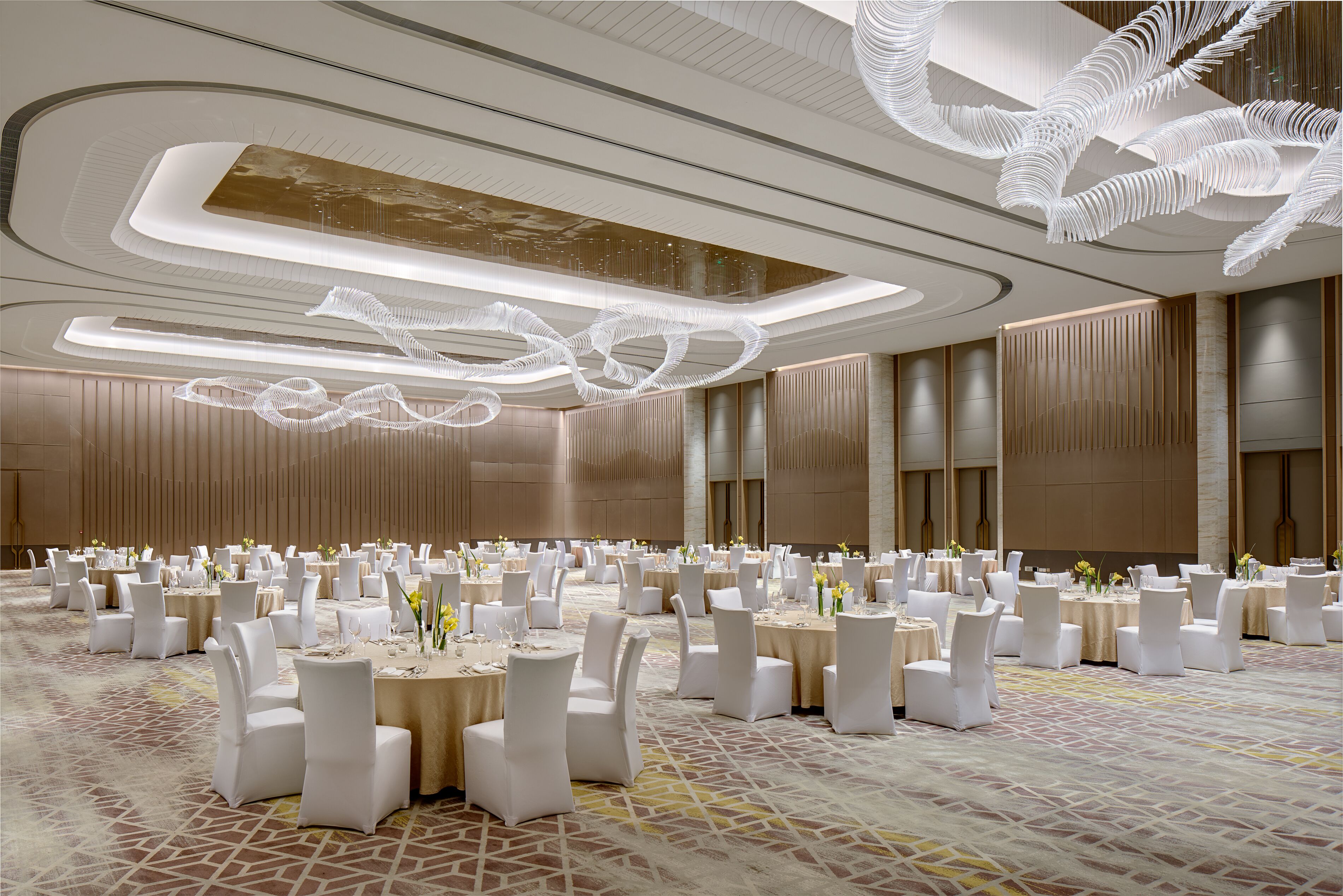A spacious banquet hall with round tables, white chairs, and a modern ceiling design.