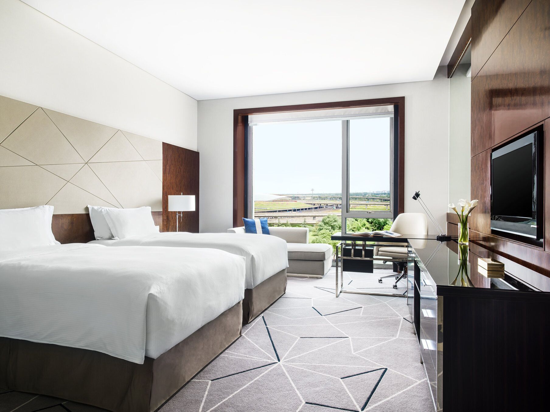 A hotel room with two beds, a desk, a TV, and a large window with a view of a sports field.