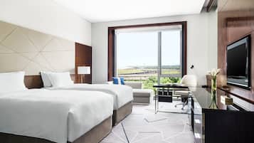 A hotel room with two beds, a desk, a TV, and a large window with a view of a sports field.