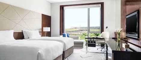 A hotel room with two beds, a desk, a TV, and a large window with a view of a sports field.