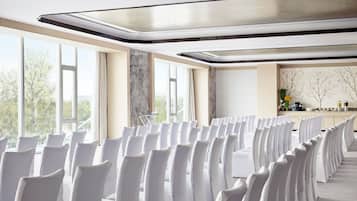 Une salle de conférence avec des rangées de chaises blanches, de grandes fenêtres et une décoration aux couleurs neutres.