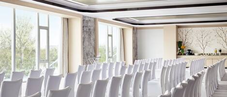 Une salle de conférence avec des rangées de chaises blanches, de grandes fenêtres et une décoration aux couleurs neutres.
