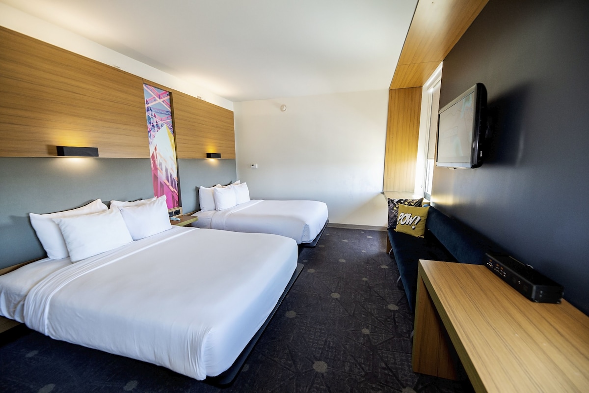 A hotel room with two beds, a TV mounted on the wall, a wooden bench, and a pillow with "POW!" on it.