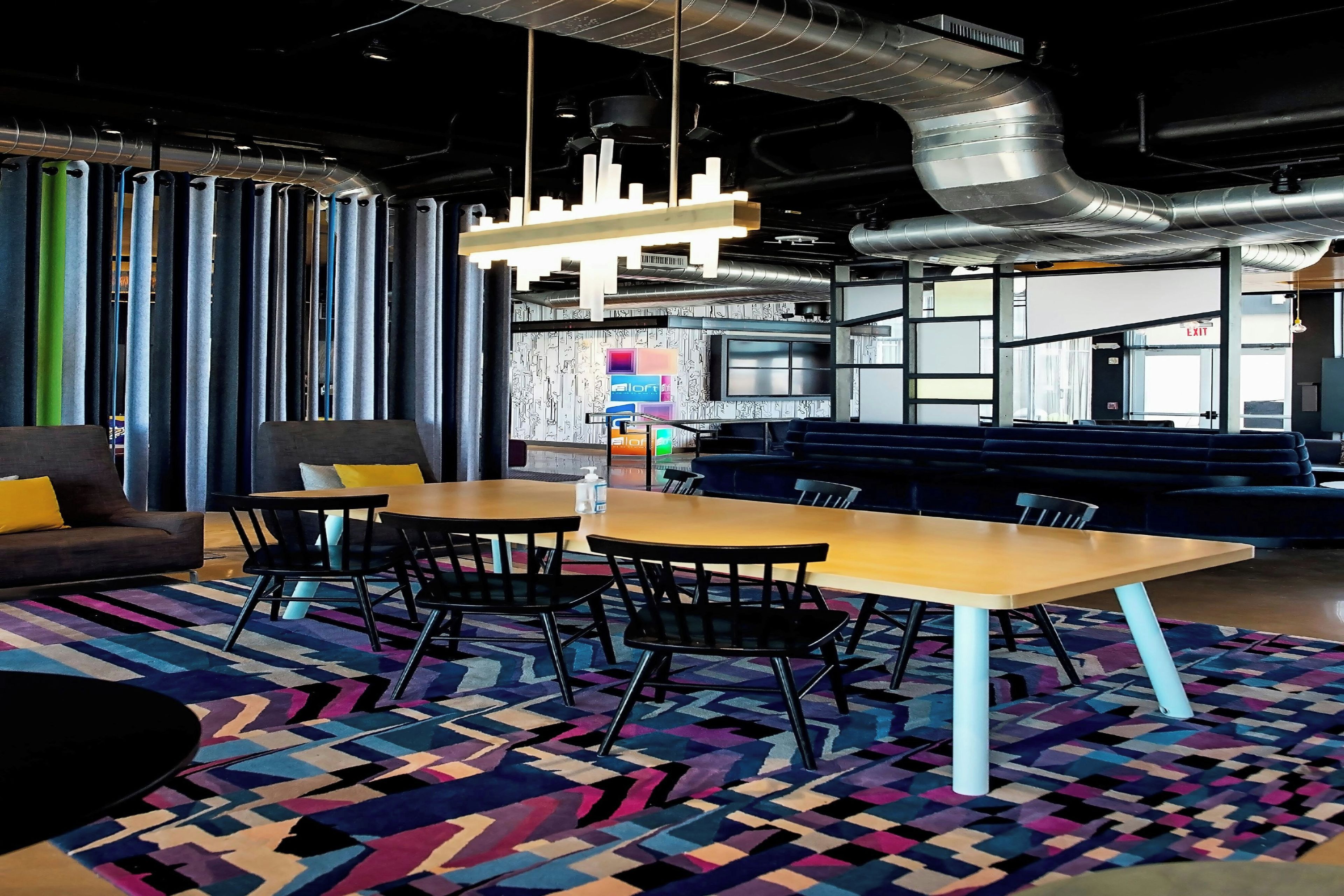 A modern lounge area with a wooden table, black chairs, and a colorful geometric rug.