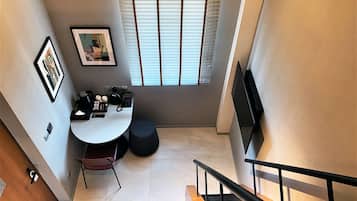 Un espacio habitable compacto con comedor, una mesita, y un sofá. Hay una ventana con persianas, un televisor de pantalla plana montado en la pared, y una escalera con barandilla.