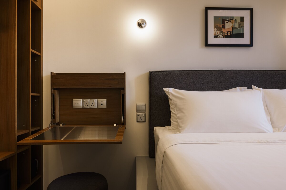 A hotel room with a bed, a small desk with a mirror, and a wall-mounted light fixture.