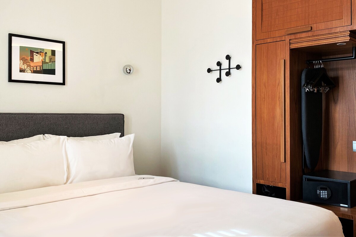 A hotel room with a bed, a coat rack, a framed picture on the wall, and a wall-mounted light fixture.