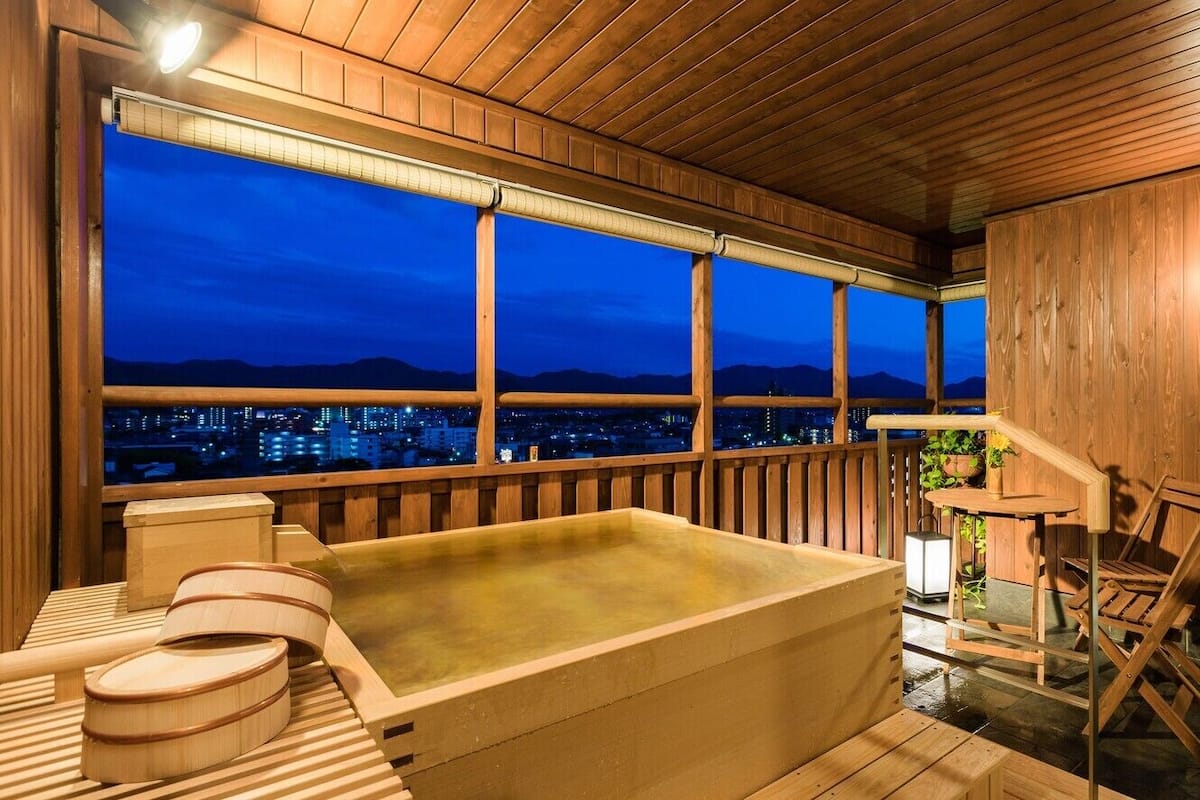Un espace de détente en bois comprenant un jacuzzi, des chaises en bois et une vue sur le paysage urbain au crépuscule.