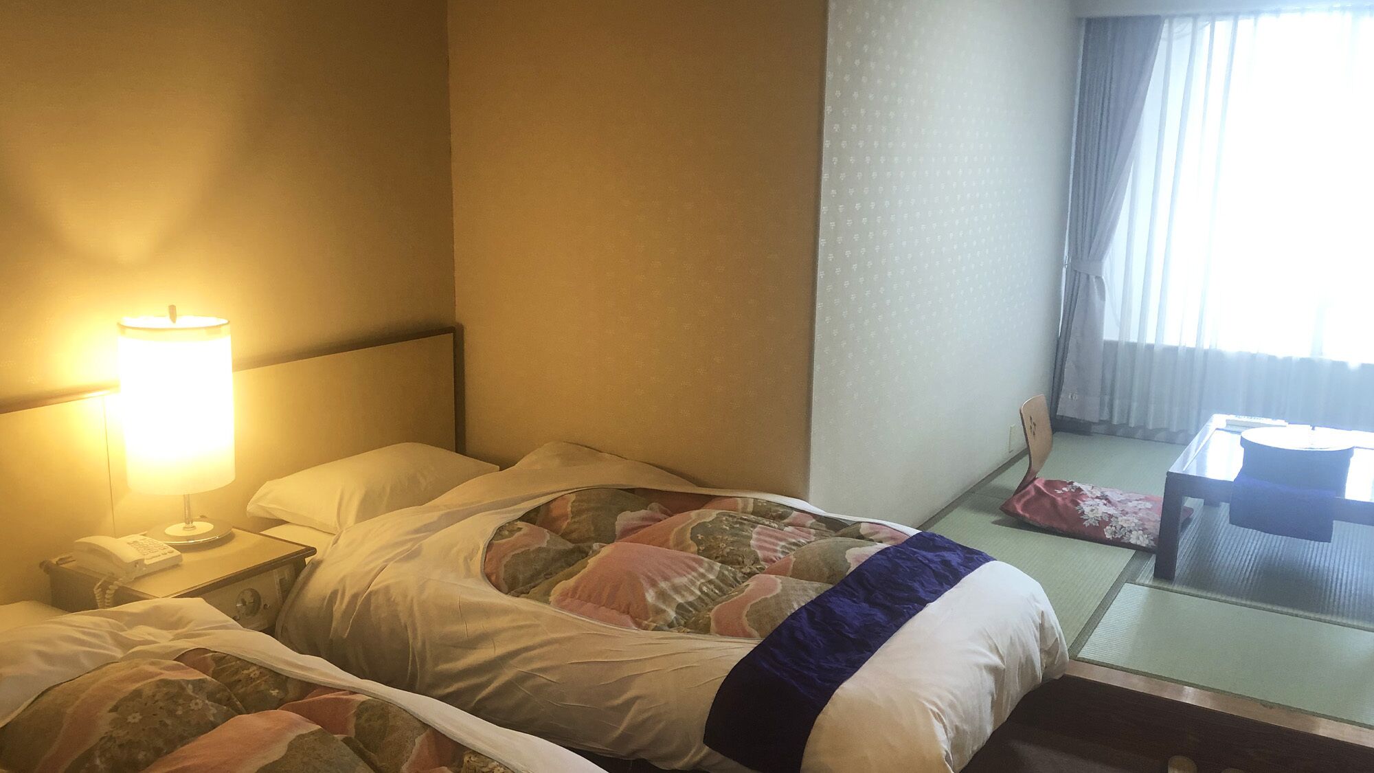 Una habitación de hotel con cama, lámpara de noche, silla y ventana con cortinas.