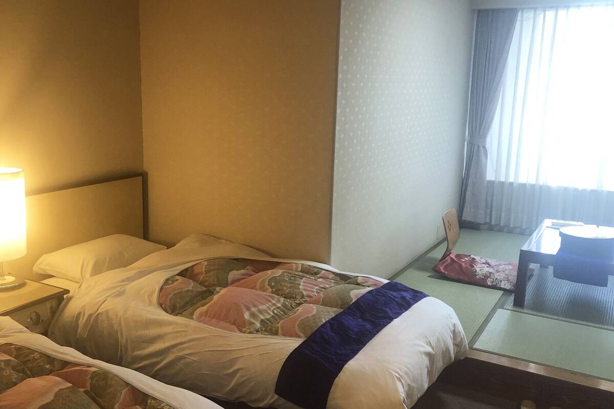 A hotel room with a bed, a bedside lamp, a chair, and a window with curtains.