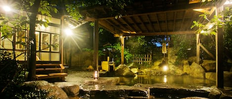 A traditional Japanese onsen with a wooden pavilion, stone bath, and a small wooden structure with steps leading to a viewing area.