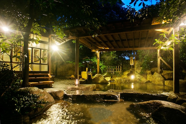 A traditional Japanese onsen with a wooden pavilion, stone bath, and a small wooden structure with steps leading to a viewing area.