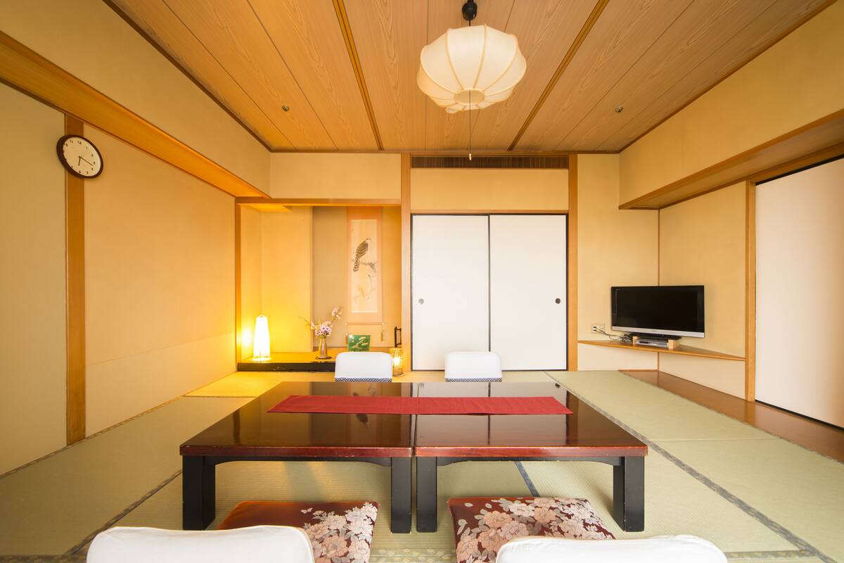 A traditional Japanese room with tatami flooring, a low wooden table, two tatami mats as seating, a television, and a shoji screen door.