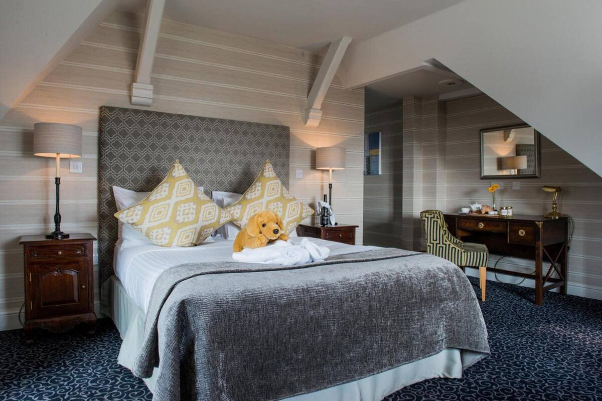 A hotel room with a large bed, a desk, and a chair. There is a teddy bear on the bed.