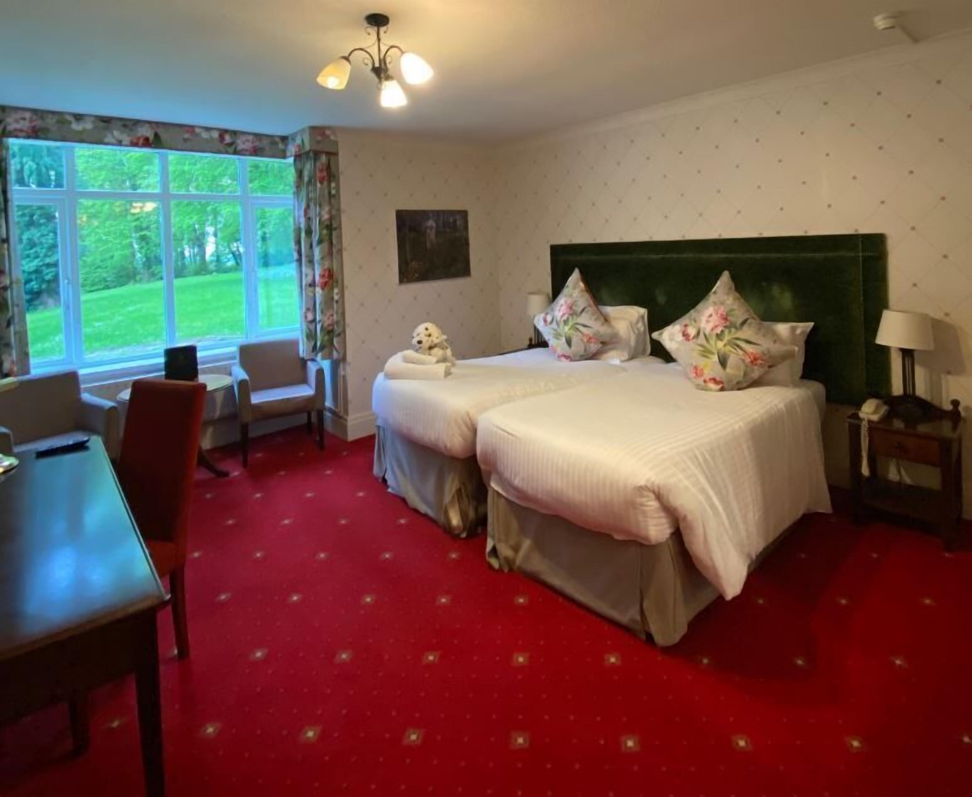 Quarto de hotel com duas camas, uma escrivaninha, uma cadeira, uma luminária e vista para o verde pela janela.