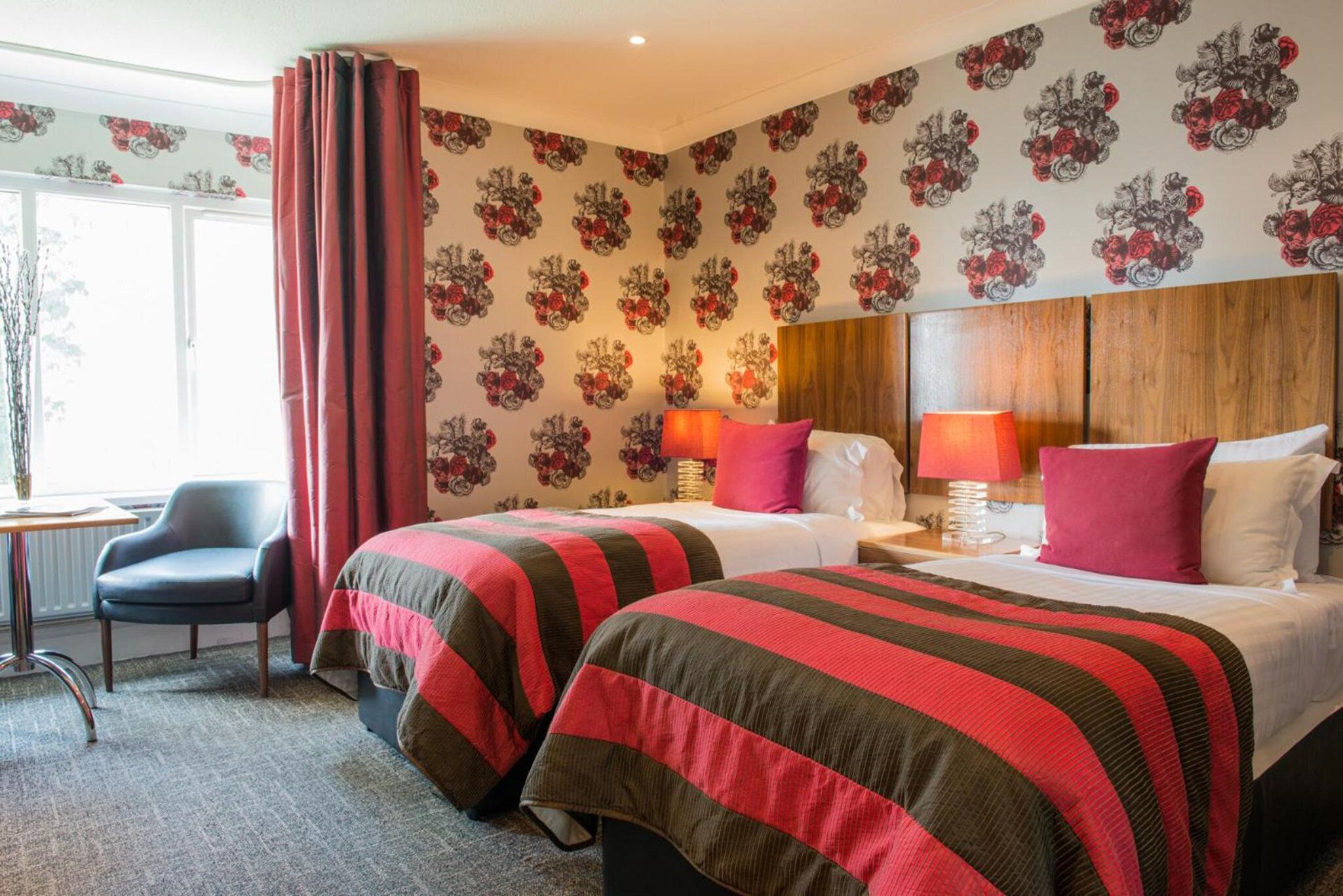 A hotel room with two beds, a wooden headboard, a nightstand with lamps, a chair, and a window with curtains.