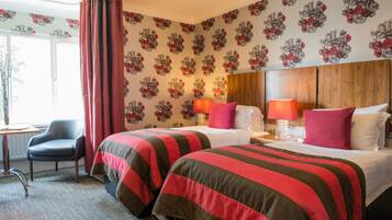 A hotel room with two beds, a wooden headboard, a nightstand with lamps, a chair, and a window with curtains.