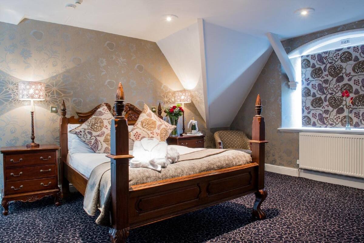 A bedroom with a four-poster bed, two bedside tables with lamps, a nightstand, a window with patterned curtains, and a radiator.