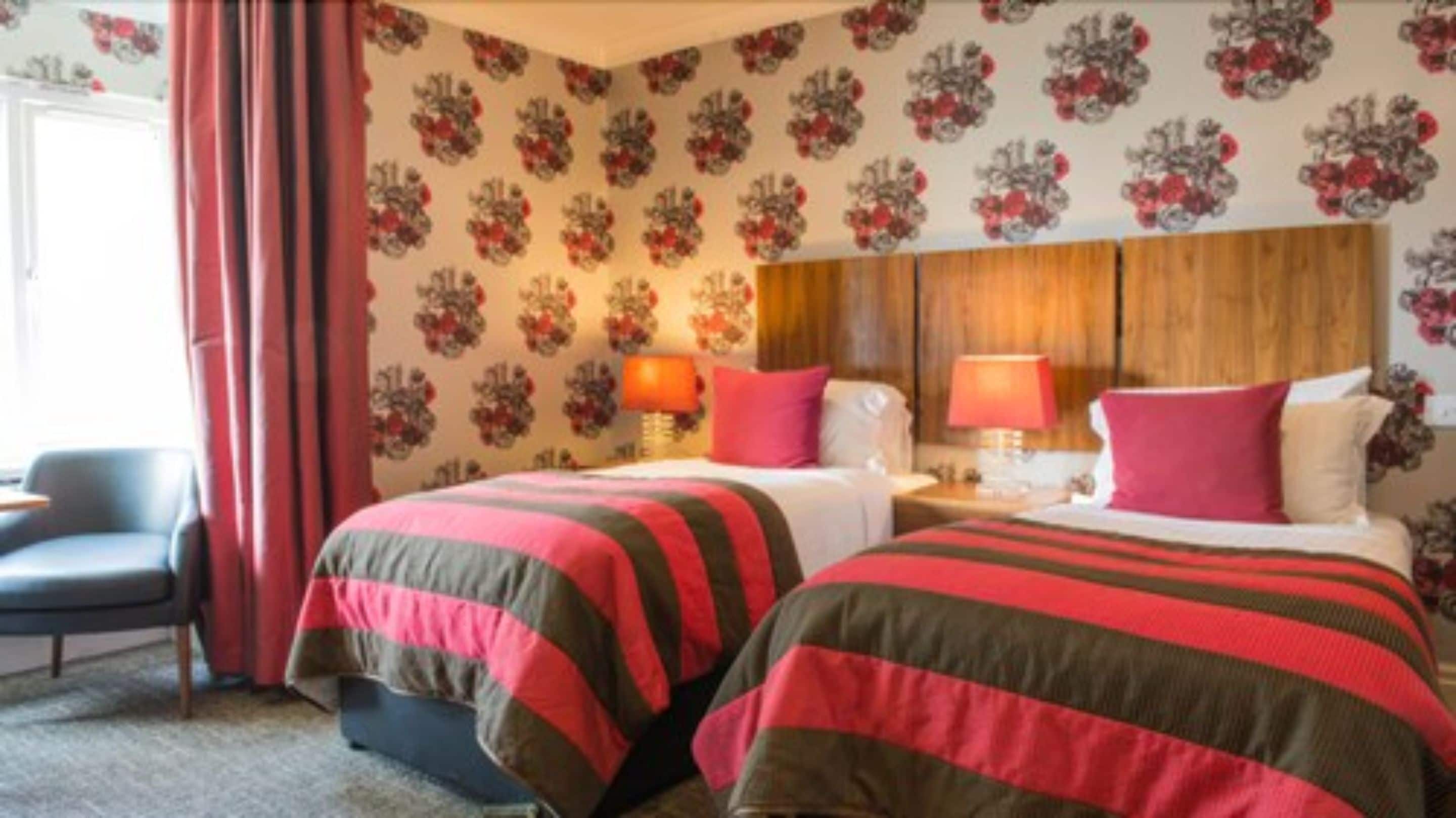 Hotelkamer met bloemenbehang, twee bedden met gestreept beddengoed, een houten hoofdbord en een bureau met een lamp.