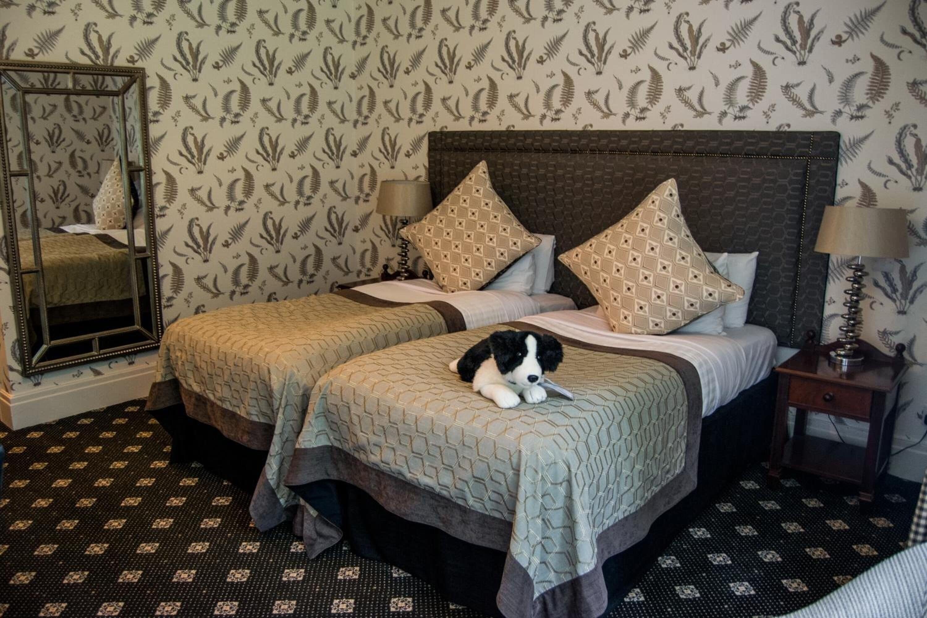 A hotel room with two beds, patterned wallpaper, a mirror, and a nightstand with a lamp.