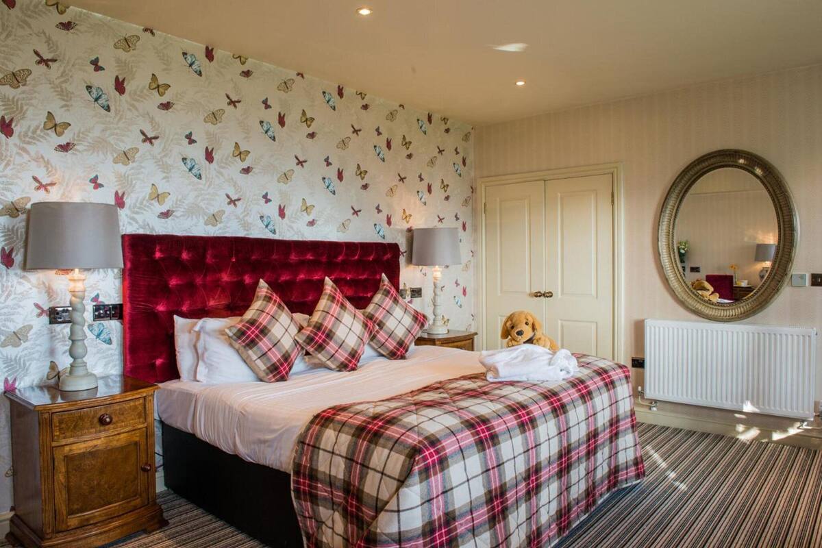 A bedroom with a red velvet headboard, a plaid bedspread, and a dog on the bed. There is a large round mirror on the wall and a radiator in the corner.