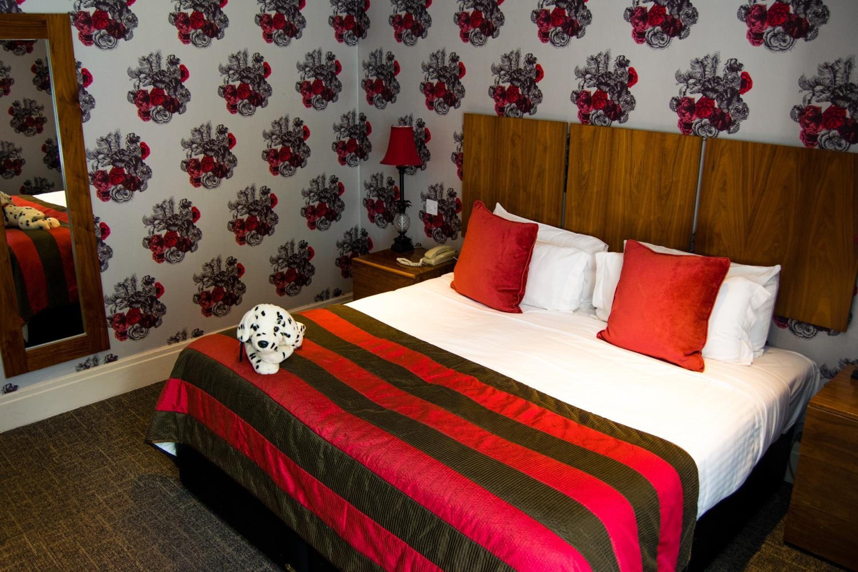Ett hotellrum med en stor säng, ett rött och svart randigt sängöverkast, ett träfärgat sänggavel och en blommig tapet.