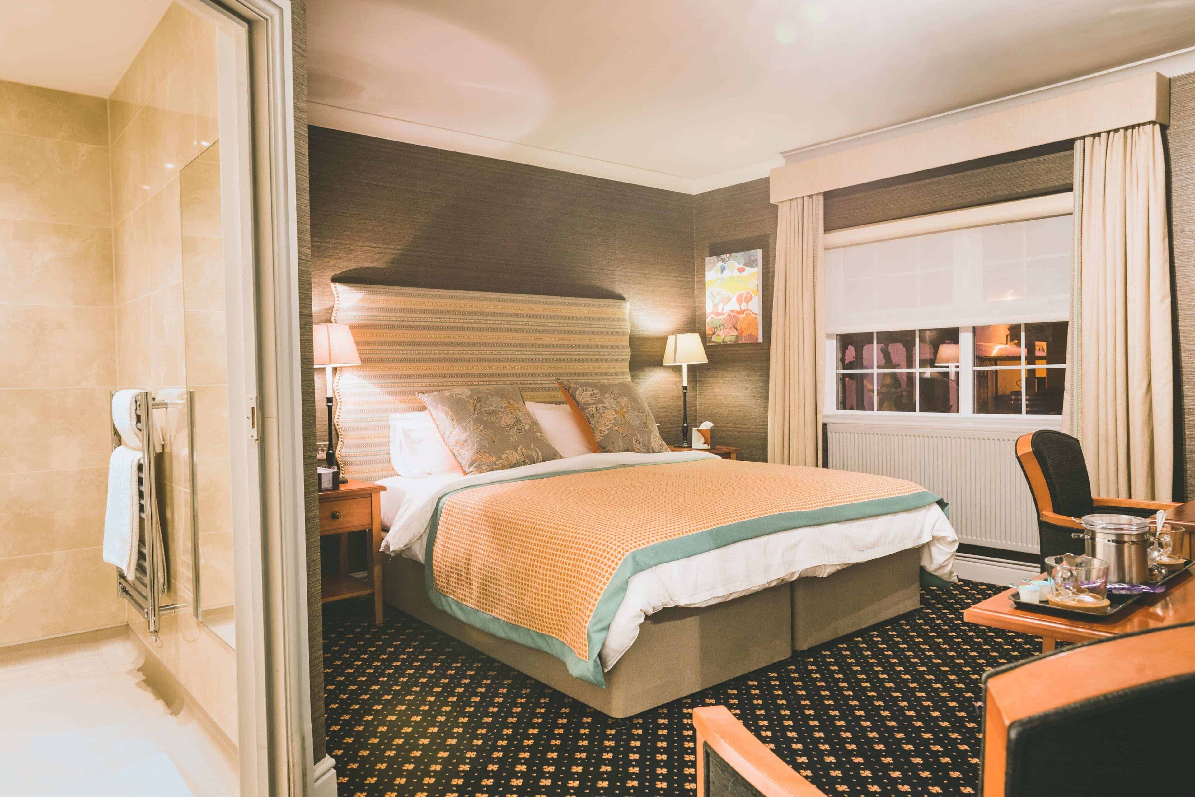 A hotel room with a large bed, a bedside table with a lamp, a window with curtains, and a view of a cityscape.