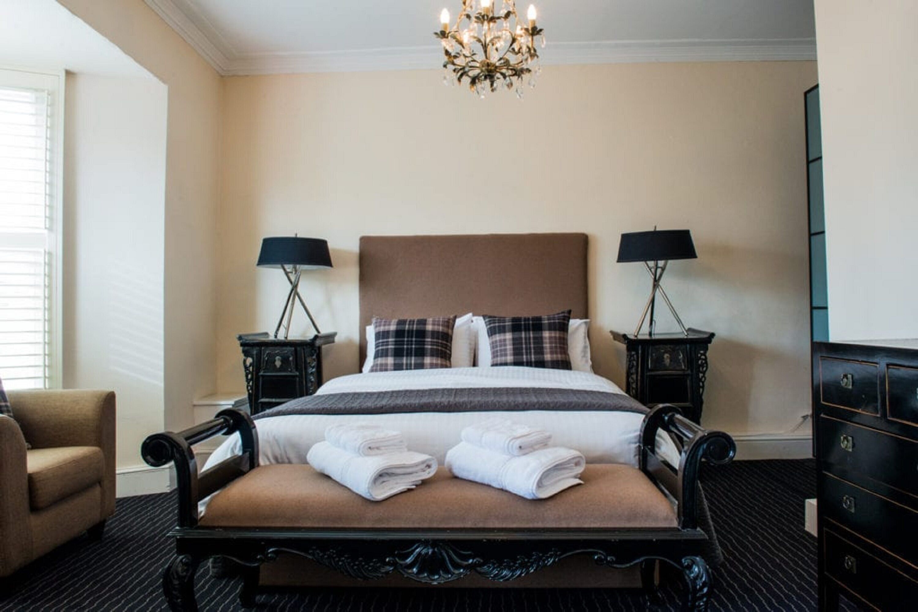 A hotel room with a large bed, two bedside tables with lamps, a chandelier, a chair, and a dresser.