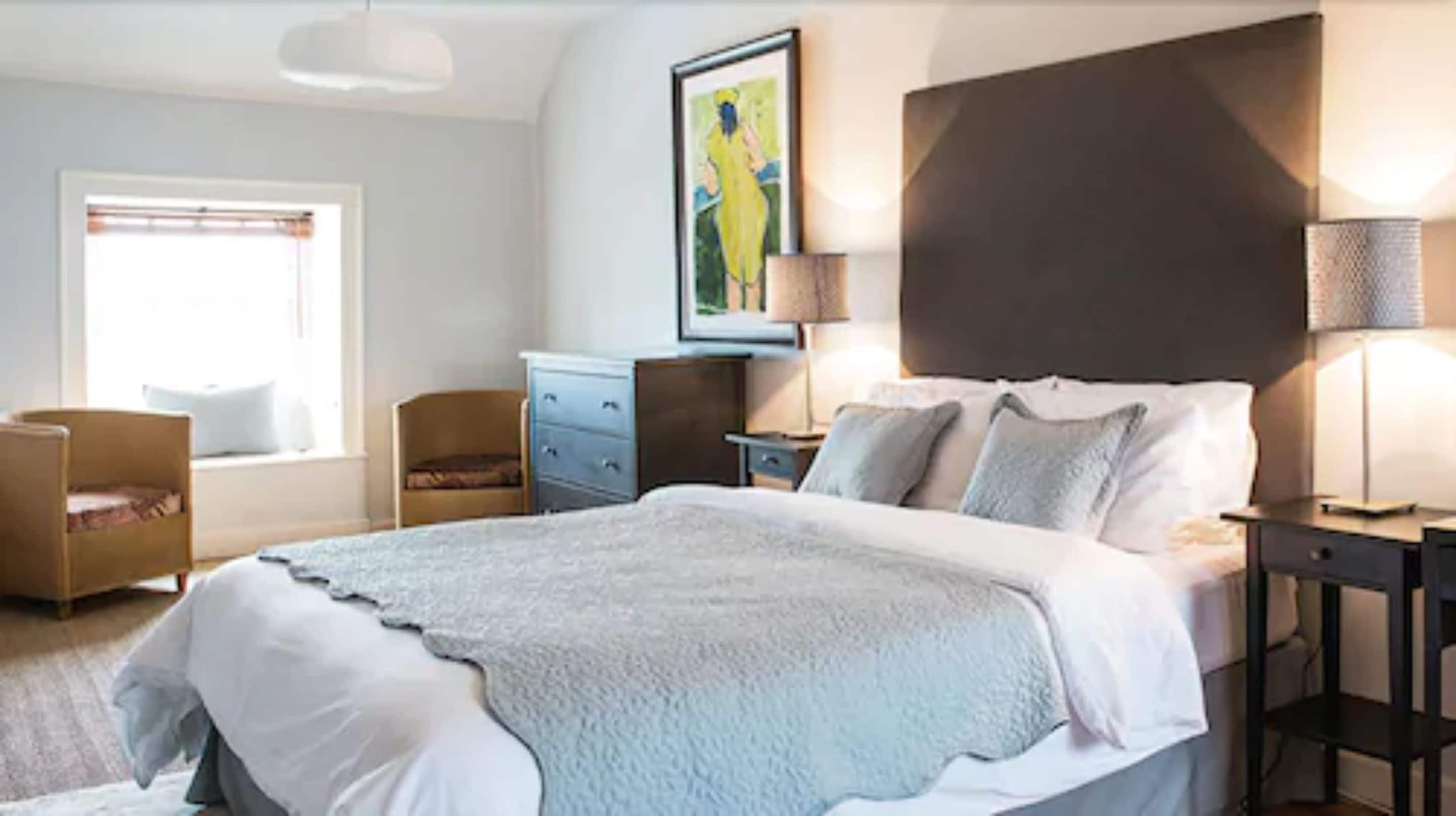 Un dormitorio con una cama grande, mesitas de noche, un aparador, una silla y una ventana con persianas.